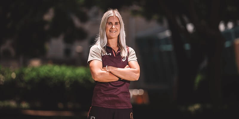 A woman with long blond hair stands outdoors with arms crossed, wearing a maroon and white sports uniform. Trees and a building are blurred in the background.