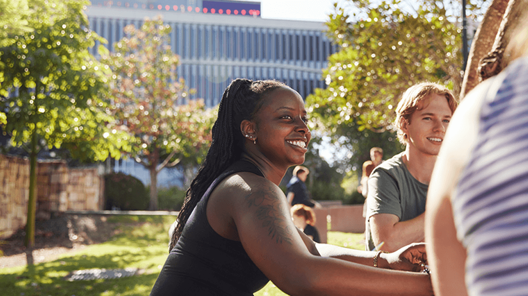 A woman and man sit outside at a picnic table, smiling and talking, with trees and a modern building in the background at UniSQ Springfield.