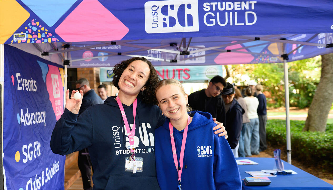 Two students wearing UniSQ Student Guild hoodies smile and pose under a Student Guild tent at an outdoor event, with banners and a table in the background.