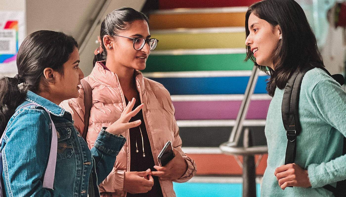 Three young women stand in conversation in front of rainbow-colored stairs; one holds a phone, and two wear backpacks.