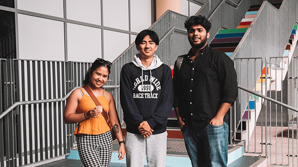 Three people stand in front of a staircase with rainbow-colored steps, posing for a photo outside a modern building.