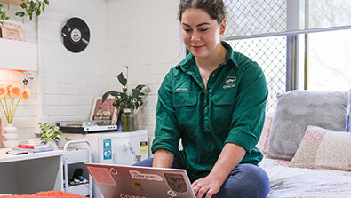 A woman sits on a bed using a laptop in a neatly decorated room with plants, a record, and a turntable visible in the background.