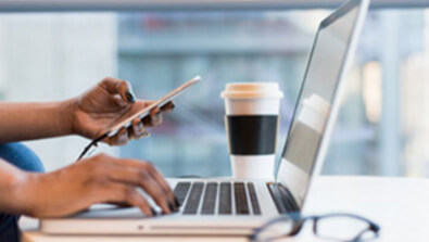 A person uses a smartphone and types on a laptop at a desk with a takeaway coffee cup in the background.