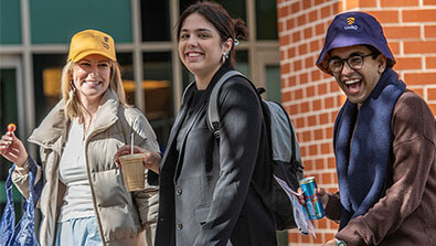 Three people stand outside, smiling. One holds a drink, another carries a backpack, and the third holds a can, with a brick wall and windows in the background.