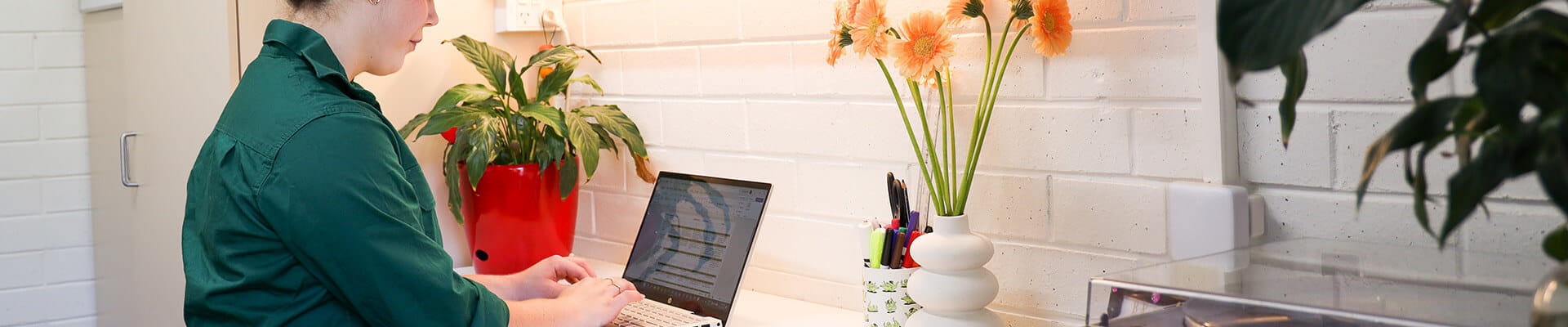 Person in a green shirt typing on a laptop at a desk with a vase of orange flowers and potted plants against a white brick wall.