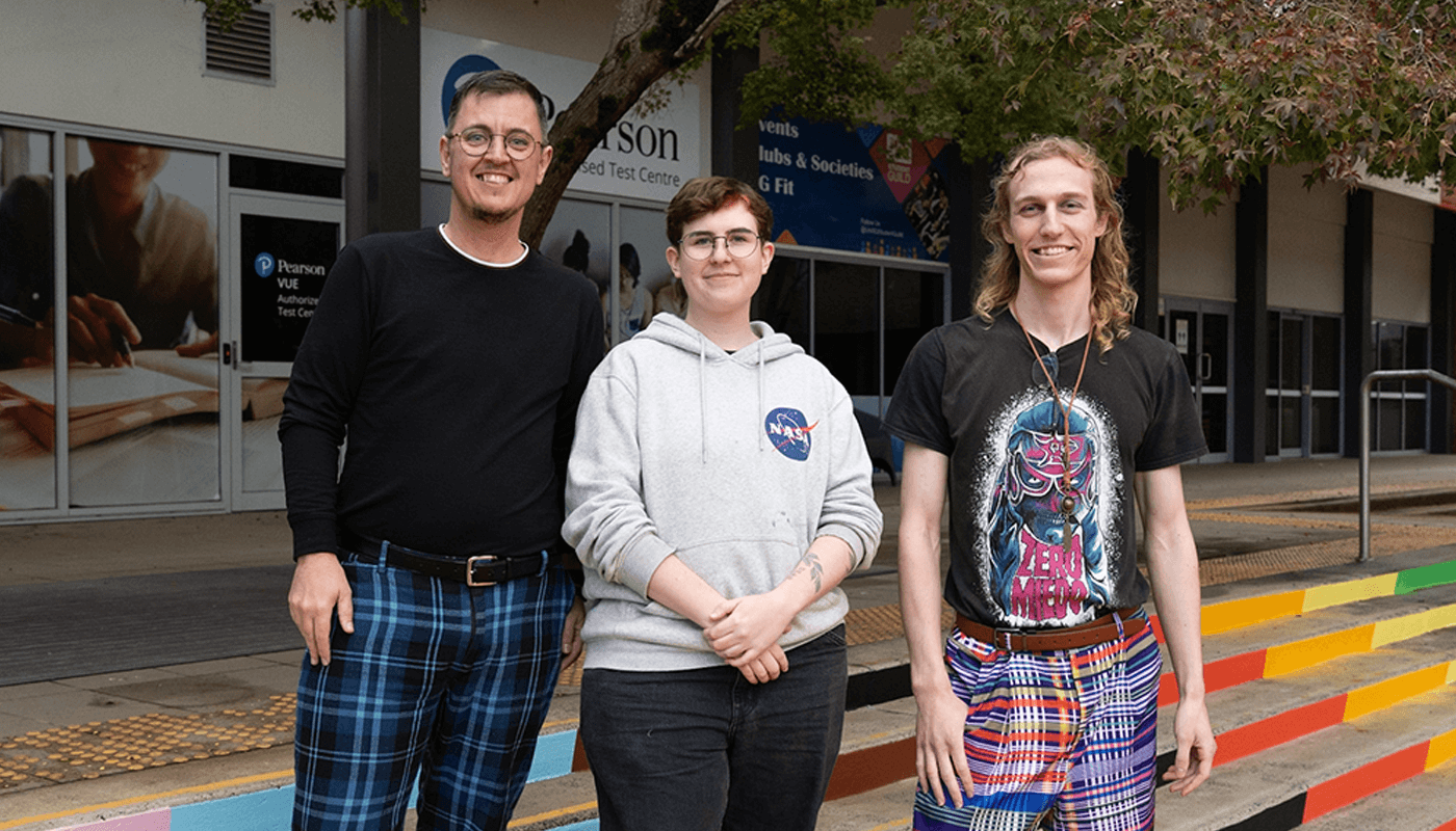 Three people stand side by side in front of a building with stairs painted in the pride colours in the background, smiling at the camera.