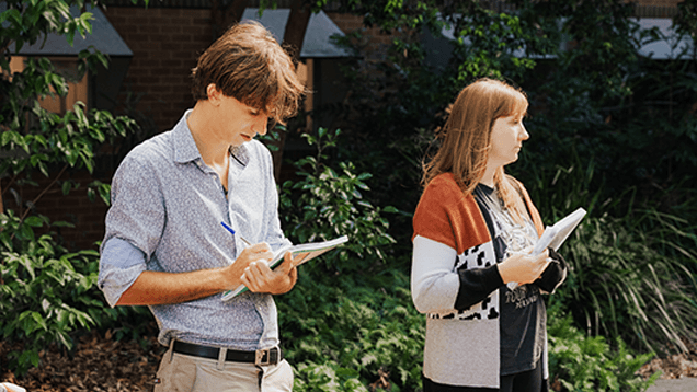 Two people standing outdoors, both holding notebooks and writing, with trees and greenery in the background.