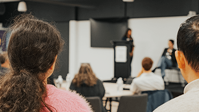Audience members watch two speakers at the front of a room during a presentation or panel discussion.