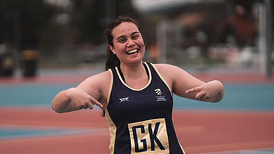 A smiling netball player in a "GK" bib points to herself on an outdoor court.