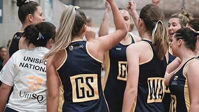 A group of female netball players in navy and gold uniforms gathers in a huddle, raising their arms in teamwork before a game.