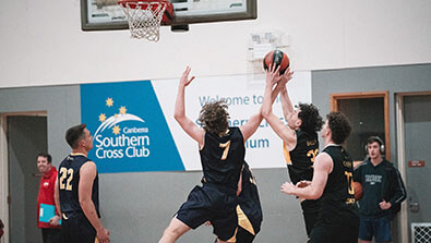 Four basketball players in dark jerseys compete near the hoop; one jumps to block a layup attempt while others watch. A Southern Cross Club sign is visible in the background.