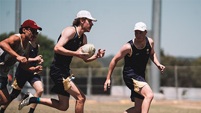 Four men in athletic gear play rugby on a grassy field, with two players in the foreground running and holding a rugby ball.