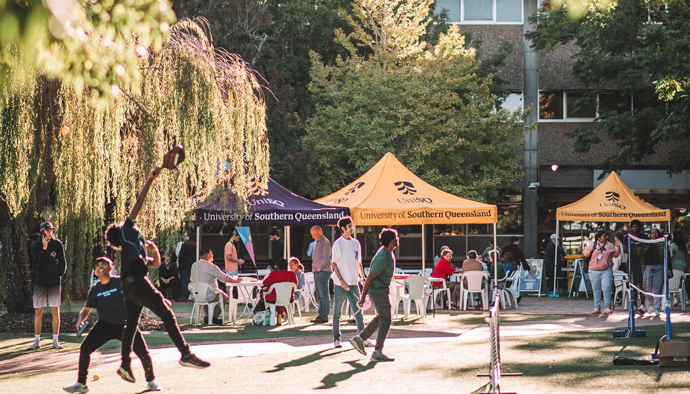 People play sports and walk on grass near University of Southern Queensland tents, with others sitting at tables under the tents in an outdoor setting.