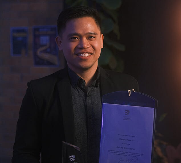 A person wearing a dark suit smiles while holding a plaque and an award certificate in a dimly lit indoor setting.
