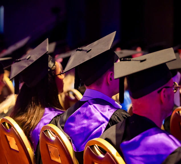Rows of graduates in caps and gowns sit on yellow chairs, facing forward during a graduation ceremony.