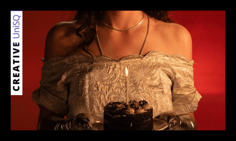 A person wearing an off-the-shoulder dress and gloves holds a small chocolate cake with a single lit candle.
