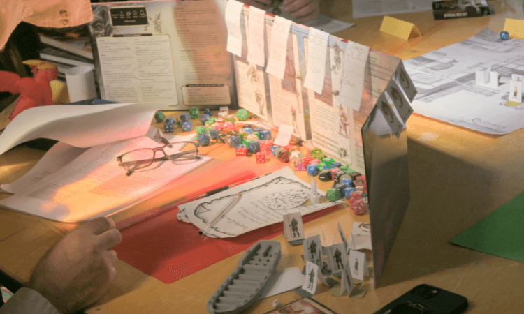 A tabletop role-playing game session in progress, with dice, character sheets, a game master screen, paper figures, and various papers spread out on a wooden table.