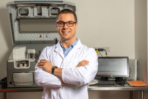 A person wearing glasses and a lab coat stands with arms crossed in front of laboratory equipment and a computer.