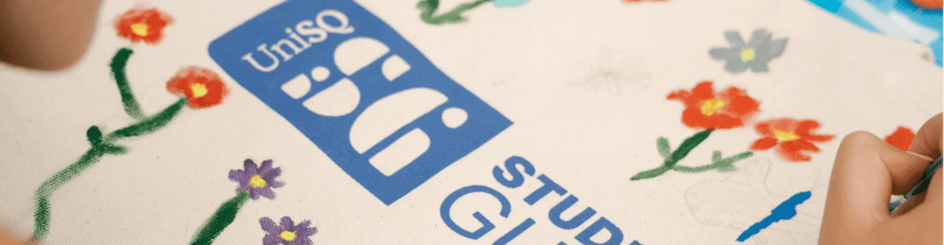 Close-up of hands painting colorful flowers around a blue UniSQ Student Guild logo on a canvas.