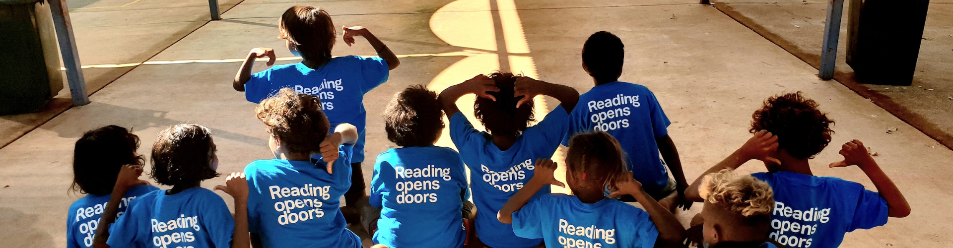 A group of children sit on the ground with their backs to the camera, wearing blue shirts that read “Reading opens doors.”.