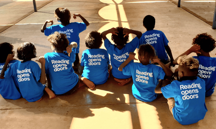 A group of children sit on the ground with their backs to the camera, wearing blue shirts that read “Reading opens doors.”.