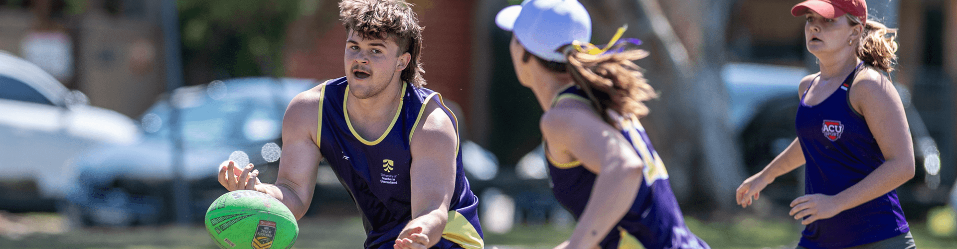 Three people playing touch football outdoors, wearing purple sports uniforms, with one player passing a green ball.
