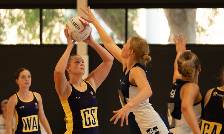 Netball players in action as a goal shooter prepares to take a shot while a defender reaches up to block.