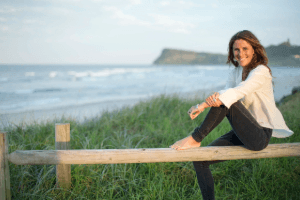 A woman sits barefoot on a wooden fence by the beach, with grass in the foreground and waves in the background under a cloudy sky.