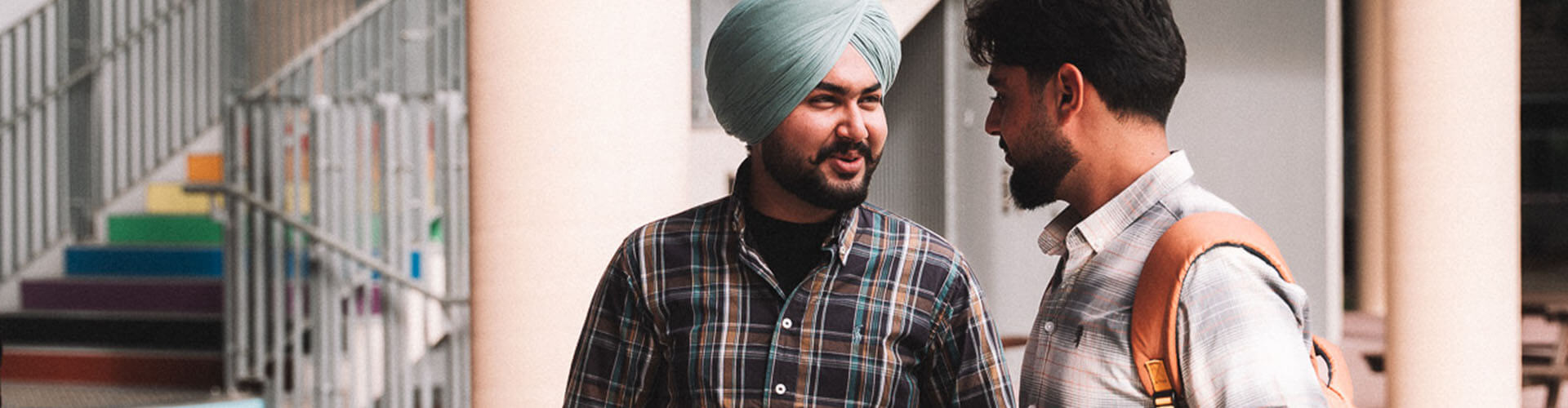 Two men stand and talk outside near a staircase with rainbow-colored steps. One man wears a turban and both are dressed casually.