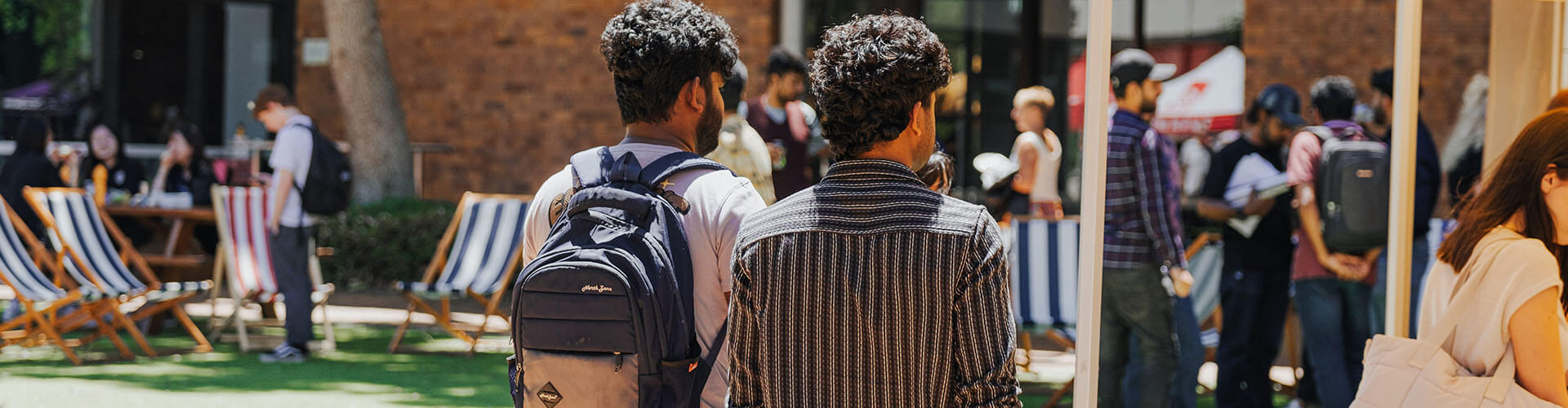 Two people with backpacks stand outdoors facing a group of people and deck chairs on a lawn, with a brick building in the background.
