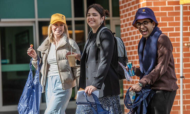 Three people walk outdoors near a brick building, holding drinks and bags, smiling and dressed in casual clothing with hats and jackets.