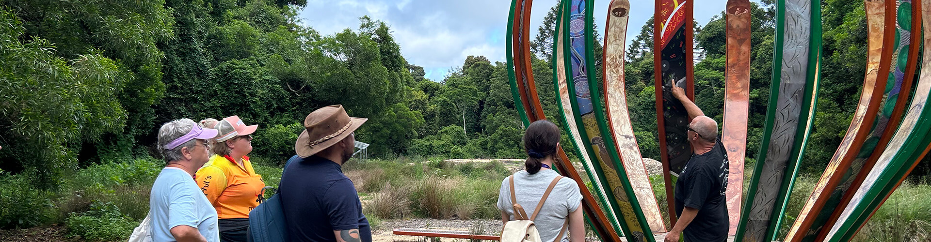 A group of people stands outdoors, observing a guide who is pointing at a colorful vertical sculpture surrounded by greenery.