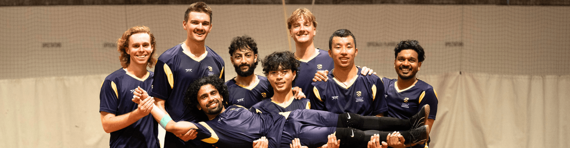 Eight men in matching navy sports uniforms pose indoors; seven stand while holding the eighth man horizontally in front of them, all smiling at the camera.