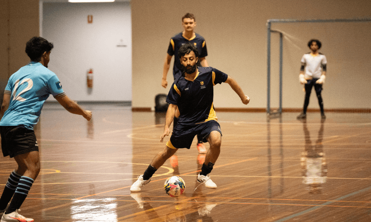 Four men play indoor soccer on a polished court; one player in dark blue controls the ball while two opponents approach and a goalkeeper stands near the goal.