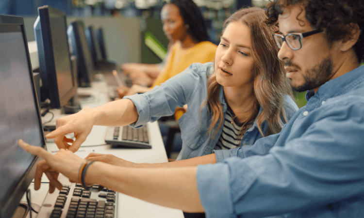 Two people working at computers, both pointing at the same screen. Another person works at a separate computer in the background.
