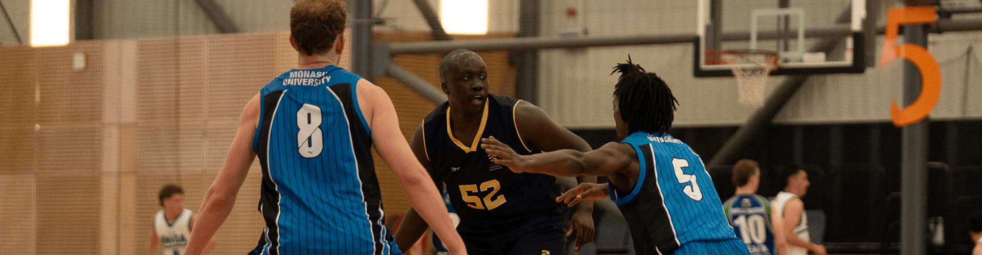 Three basketball players are on the court mid-game; one in a dark jersey with number 52 is dribbling, while two players in blue jerseys numbered 8 and 5 are defending.
