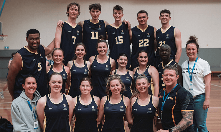 A group of basketball players and staff pose for a team photo indoors, wearing matching dark uniforms with numbers and logos.