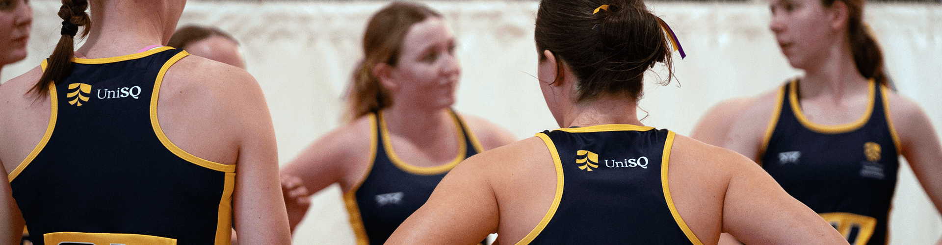 A group of female athletes in blue and yellow UniSQ uniforms stand in a circle, appearing to be in discussion during an indoor sports event.