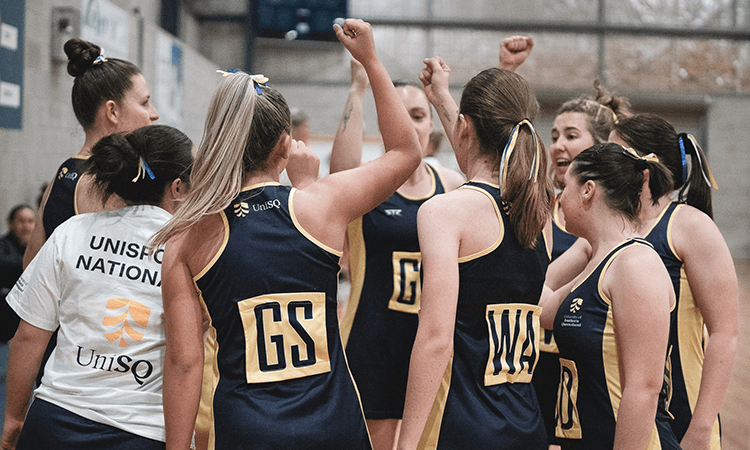 A group of female netball players in navy and yellow uniforms huddle together, raising their fists in team spirit inside an indoor court.