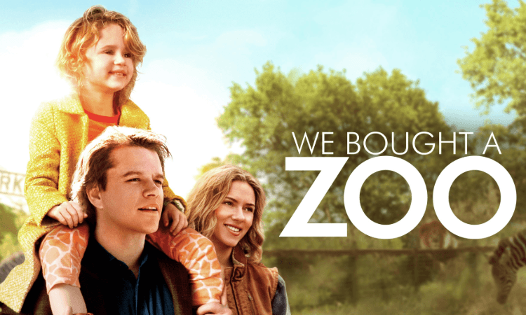A man carries a small child on his shoulders while a woman stands beside them, all smiling outdoors. The text reads, “We Bought a Zoo.”.