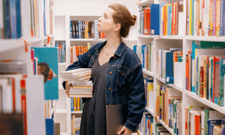 Woman in a denim jacket holding a stack of books and a laptop while browsing shelves in a brightly lit library.