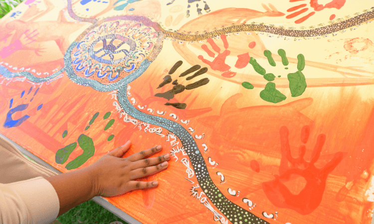 A person places their hand on a large, colorful artwork featuring painted handprints and intricate patterns on an orange background.