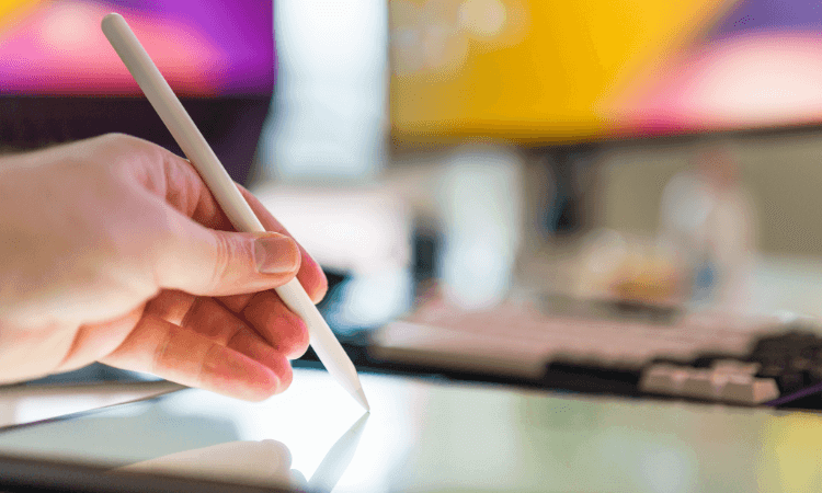A hand holds a white stylus, writing or drawing on a tablet screen, with a computer keyboard and blurred monitors in the background.
