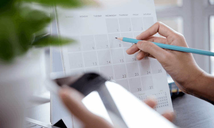 A person holding a pencil points to a date on a desk calendar while using a smartphone with their other hand.