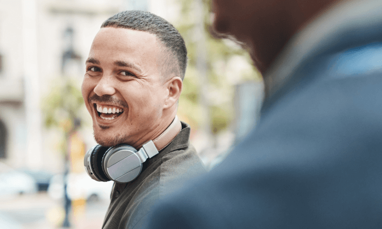A man with short hair and headphones around his neck smiles while standing outdoors, with another person blurred in the foreground.