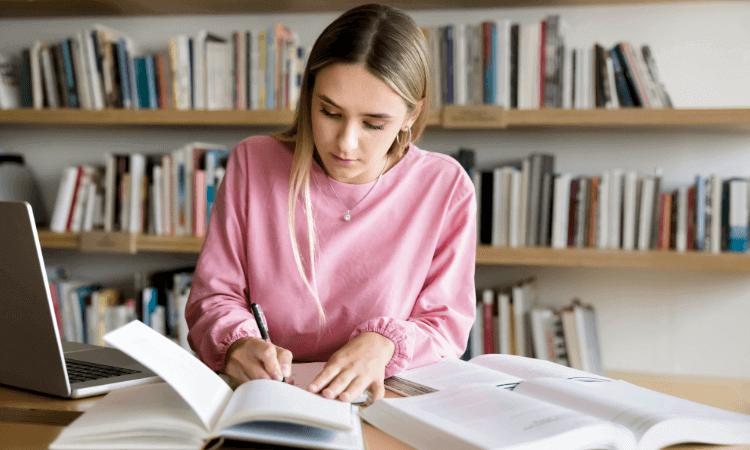A woman sits at a table in a library, writing in a notebook with open books and a laptop in front of her.