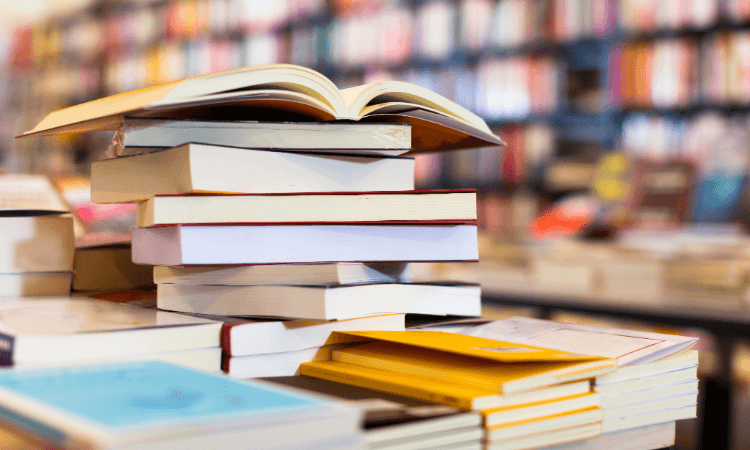 A stack of books with an open book on top sits on a table in a bookstore, with shelves of more books blurred in the background.