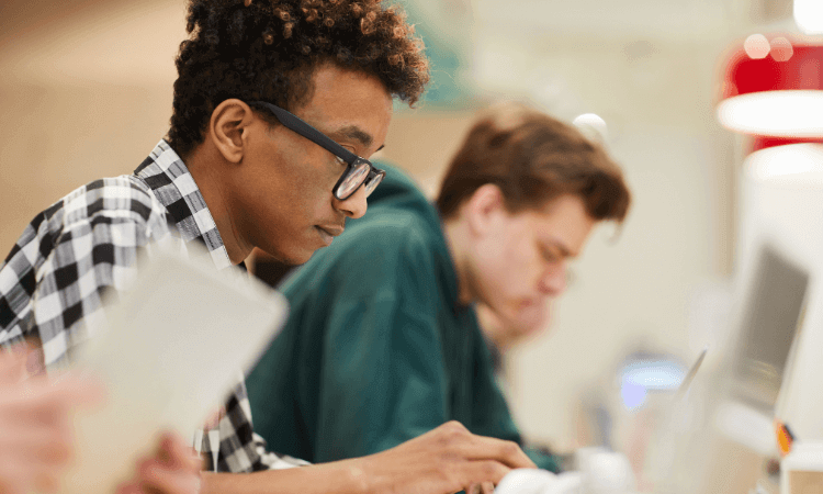 Two young people are seated indoors, working on laptops or tablets. They appear focused and are dressed casually. The background is slightly blurred.
