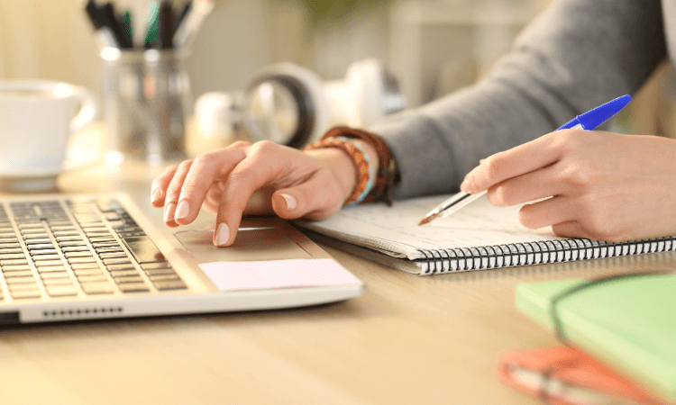 Person working at a desk with a laptop, taking notes in a spiral notebook with a pen, and surrounded by office supplies.