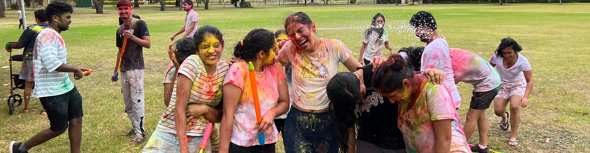 A group of people covered in colored powder celebrate outdoors on a grassy field, with some spraying water and others laughing and embracing.